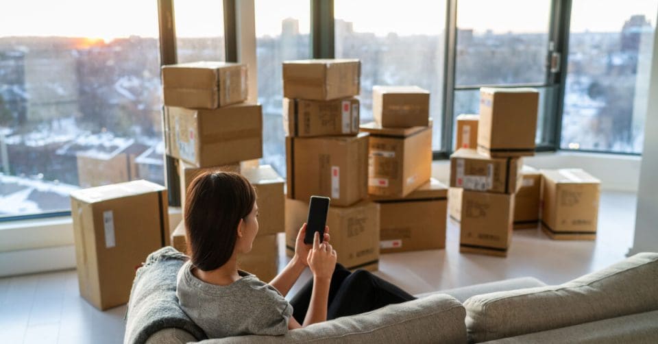 Home move out of apartment moving boxes woman using online movers services on mobile phone app easy pick-up with packages for new home. Asian new homeowner girl happy sitting in sofa.
